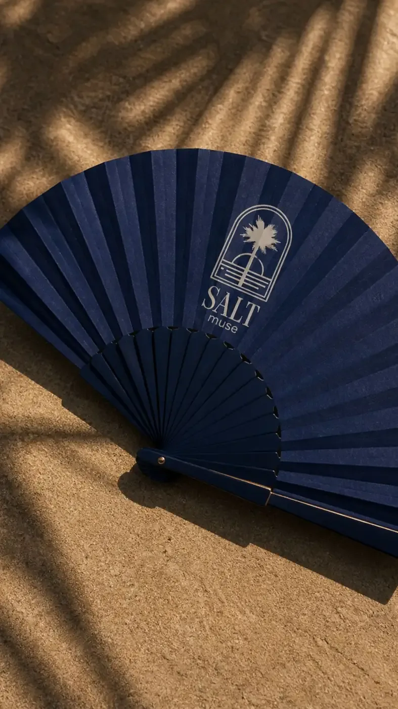 Midnight navy Salt Muse hand fan on warm sand with palm shadows